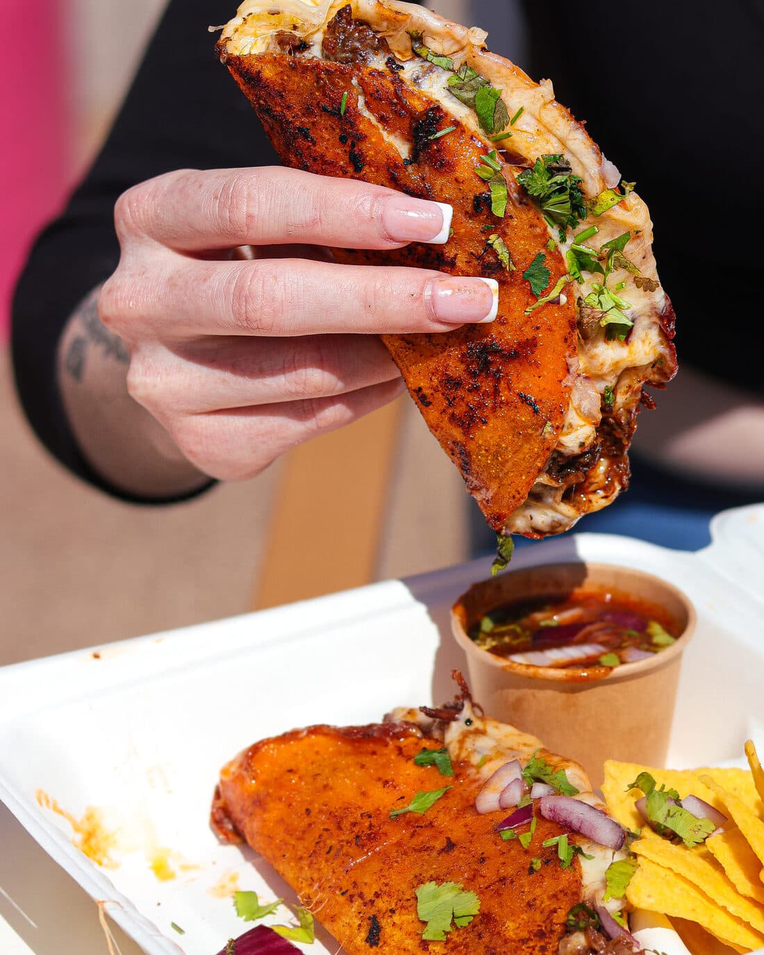 A cheesy birria quesadilla held over a tray of consommé and nachos